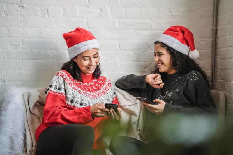 Two friends in Santa hats on a sofa, each looking at a phone