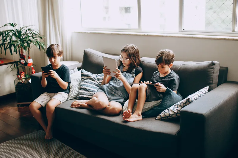 Three children side by side on a sofa, each absorbed in a phone or tablet
