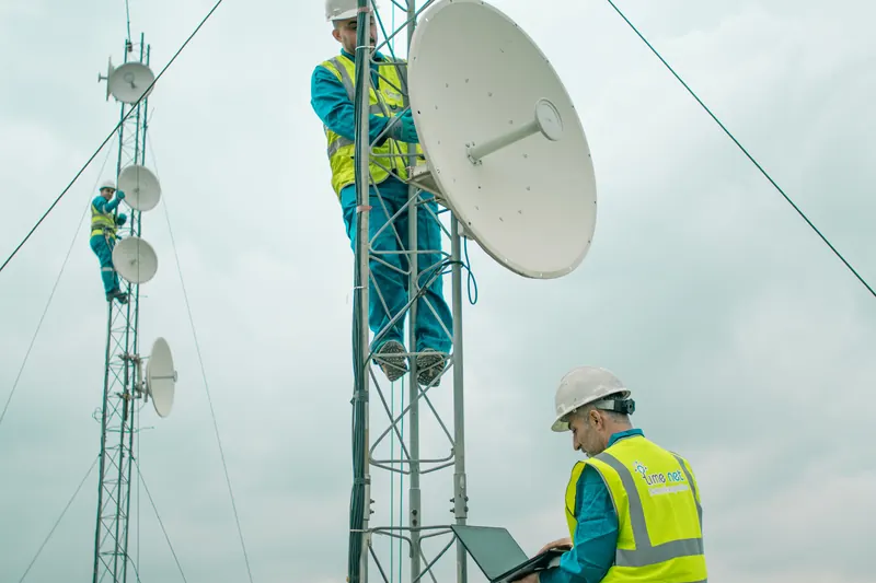 Two technicians in high-visibility vests on a rooftop, one aligning a microwave dish on a mast while the other checks a laptop